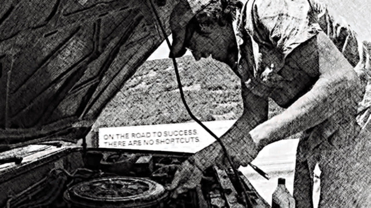 Black and white image of Steve under the hood of his broken down car, while a semi zooms by in the other direction with a sign that says "on the road to success there are no shortcuts."