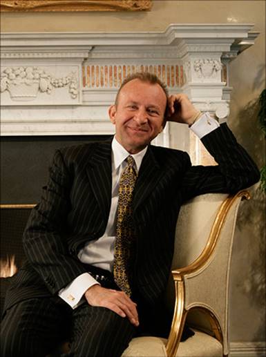 Picture of John in a suit sitting on a fancy chair in a beautiful house that he designed.